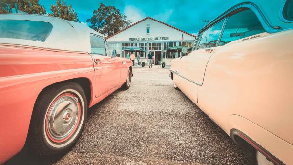 Blick zwischen zwei parkenden US-Cars hindurch auf das Merks Motor Museum im Hintergund des Bildes
