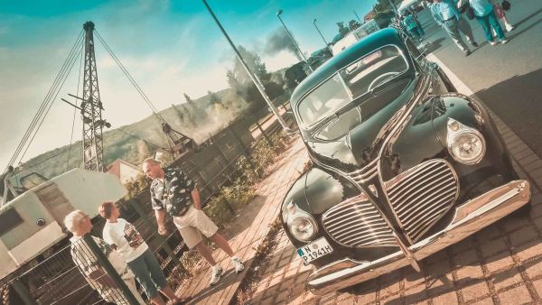 Schwarzer US-Oldtimer steht vor Gleisen. Im Hintergrund fährt eine Dampflock weg.