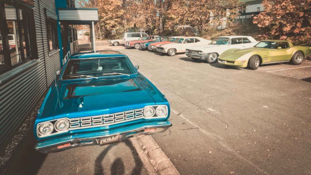 Frontansicht eines blauen Plymouth US-Oldtimer der an einer Hauswand geparkt steht. Im Hintergrund sind weitere geparkte US-Cars zu sehen.