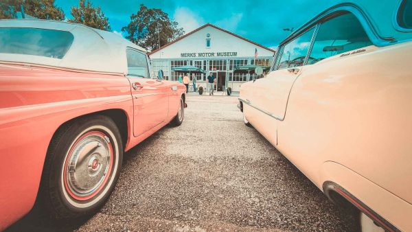 Blick zwischen zwei parkenden US-Cars hindurch auf das Merks Motor Museum im Hintergund des Bildes