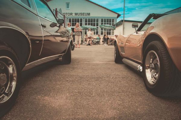 Blick zwischen zwei parkenden US-Cars hindurch auf das Merks Motor Museum im Hintergund des Bildes